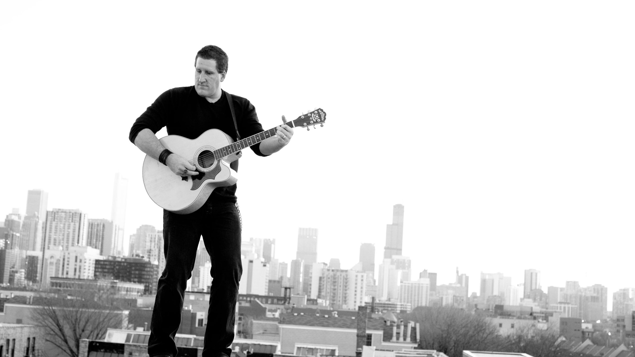 Scott DuBose playing acoustic guitar on a rooftop with the Chicago skyline behind him